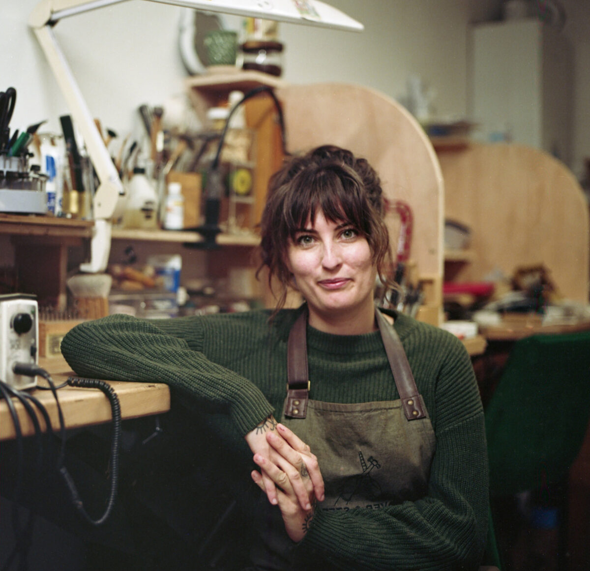 Jeweller wearing a Silver & Steel branded apron sits at a jewellery bench in a professional jewellery studio room with other benches in the background. There are tools behind her.