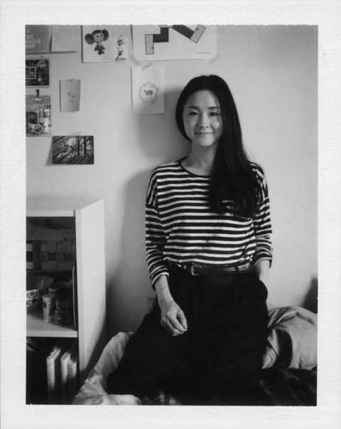 A black and white photo of a woman in an art studio. She wears a stripey long sleeved top and has long black hair