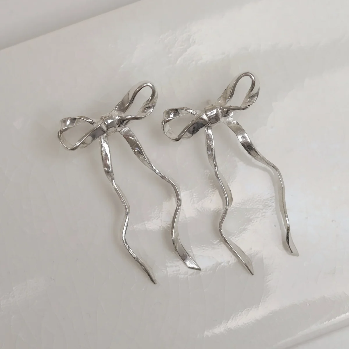 A pair of silver bow earrings rest on a white texture with light and shadow falling over them