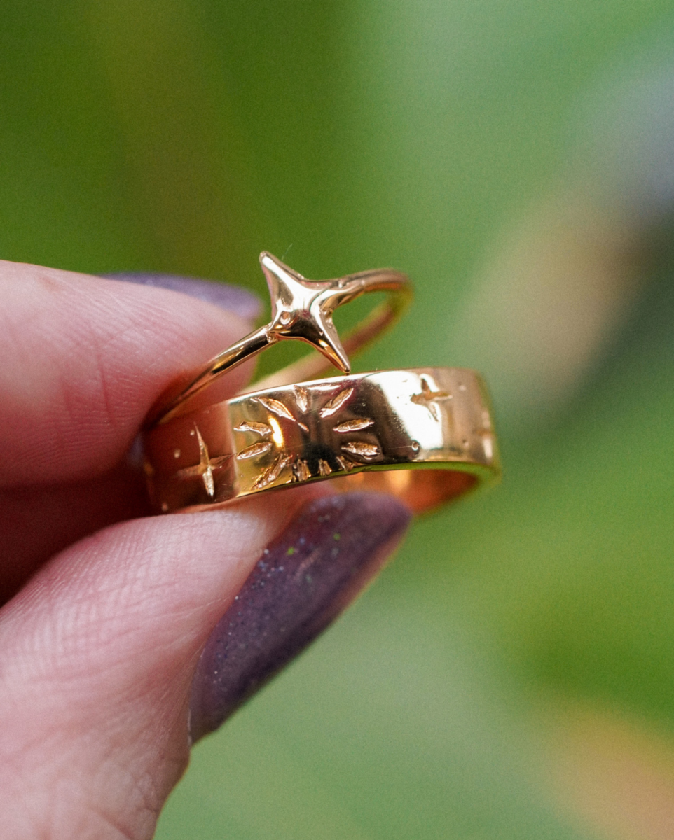 fingers with purple nails hold two gold rings. One ring is slim with a star shape and the other is a wide band with engravings that look like a sun