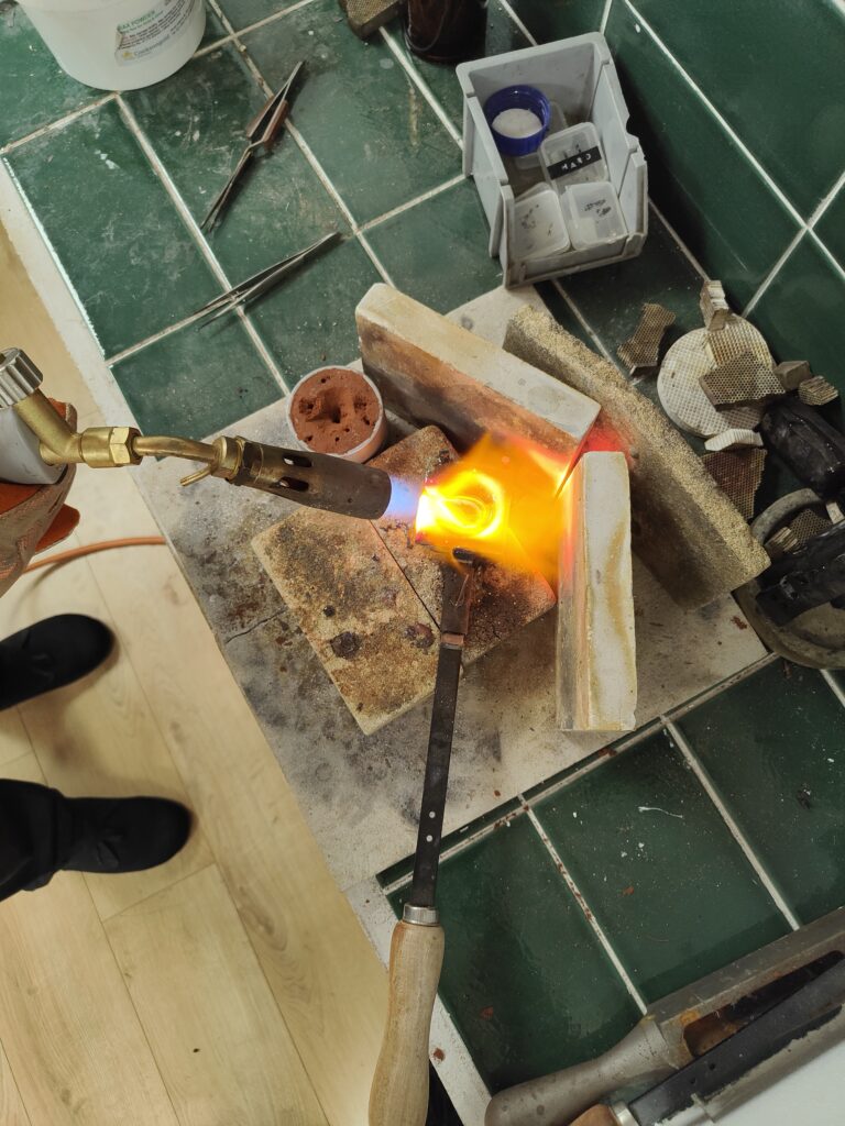 the camera looks down on a large blowtorch melting silver in a crucible for sandcasting. The delft clay mould is next to it and there are the silversmiths feet in the frame