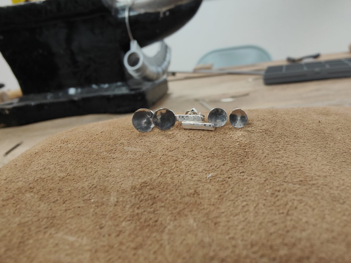 three pairs of stud earrings rest on a brown sandbag with an anvil behind