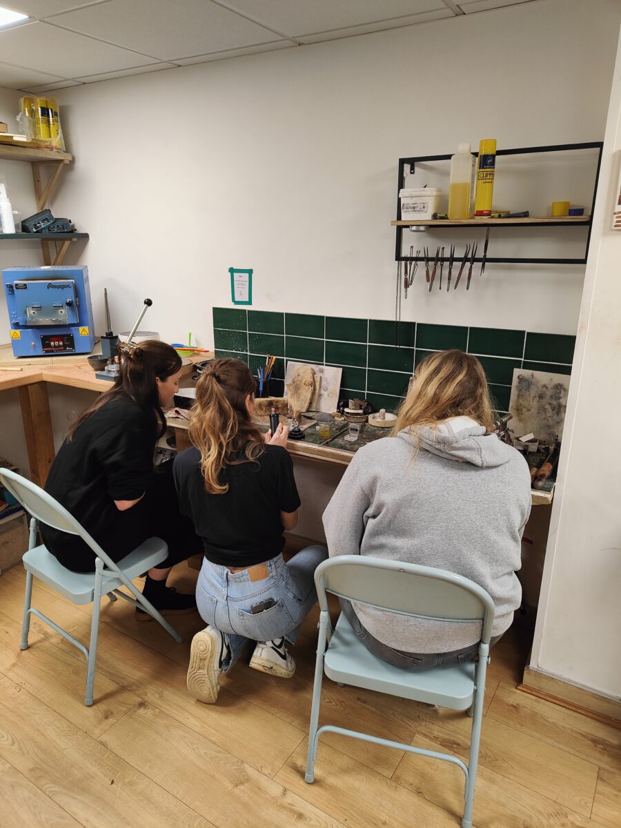 Three women in a jewellery studio gather around a soldering area to learn how to solder jewellery.