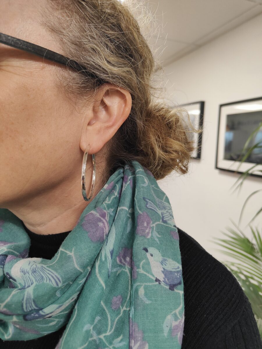 person wearing glasses and a turquoise scarf is wearing handmade silver hoop earrings that she has made herself in a workshop. There is gallery photography in the bakground.