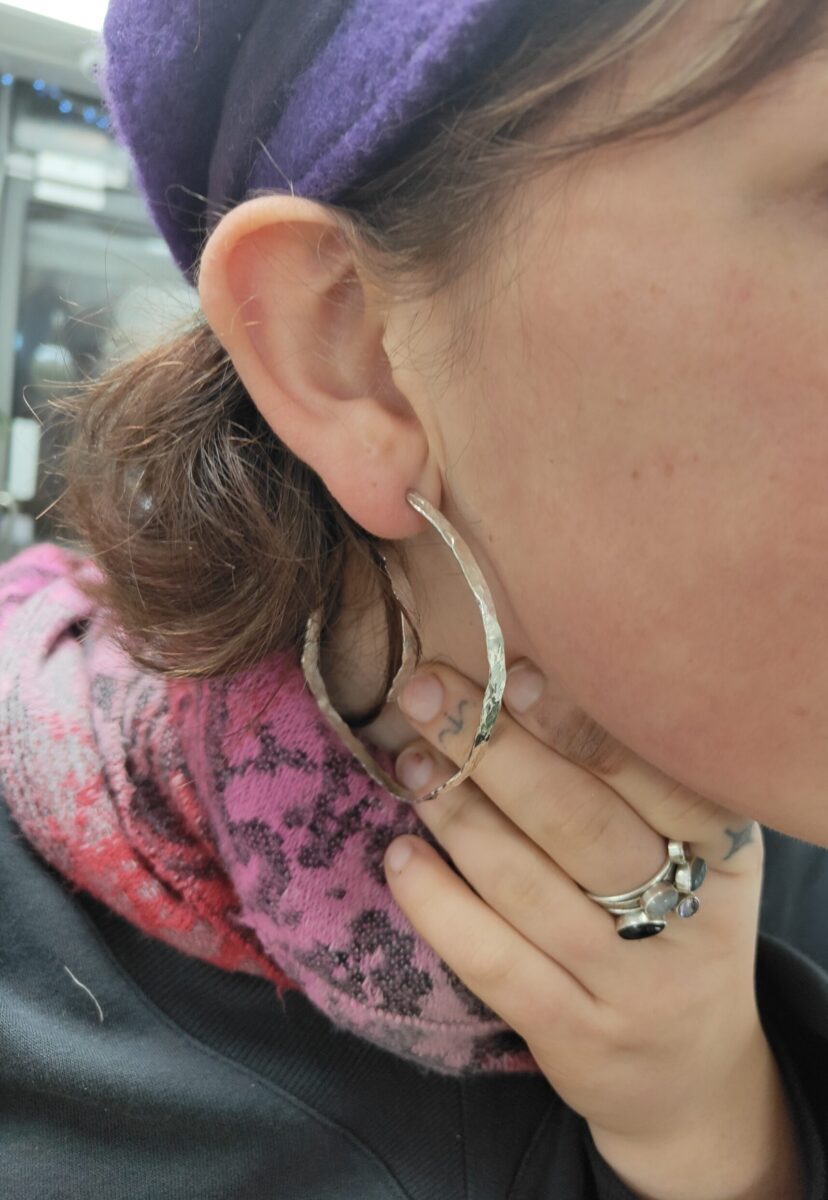 person wears textured handmade silver hoop earrings and a pink patterned scarf. They are holding the hoop to the camera with a hand wearing silver rings.