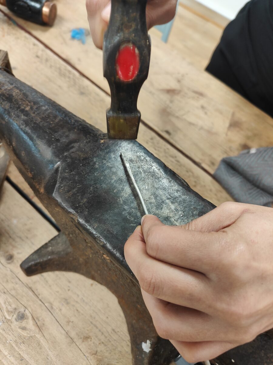 A hammer hits a piece of silver wire on an anvil in a jewellery workshop