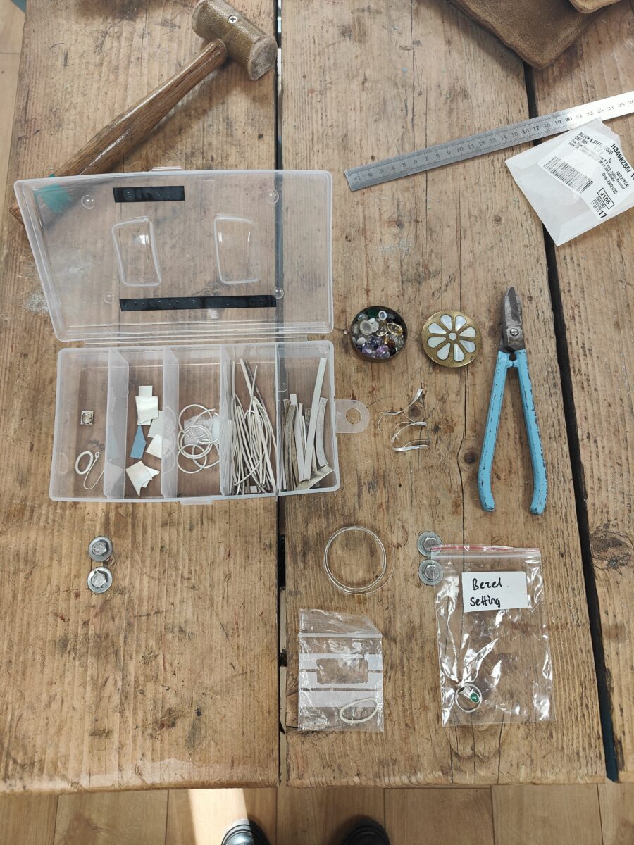 sterling silver, a selection of gemstones in a small brass tin and a pair of pliers are laid out wiht plastic bags and bezel strip on a jewellery bench
