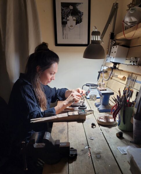 A jewellery sits at a busy jewellery bench surrounded by tools. she is working on something by lamplight.