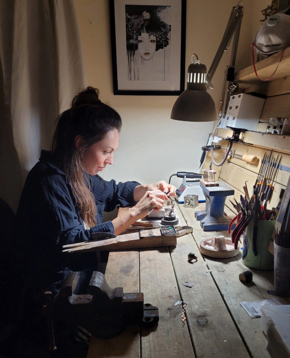 A jewellery sits at a busy jewellery bench surrounded by tools. she is working on something by lamplight.