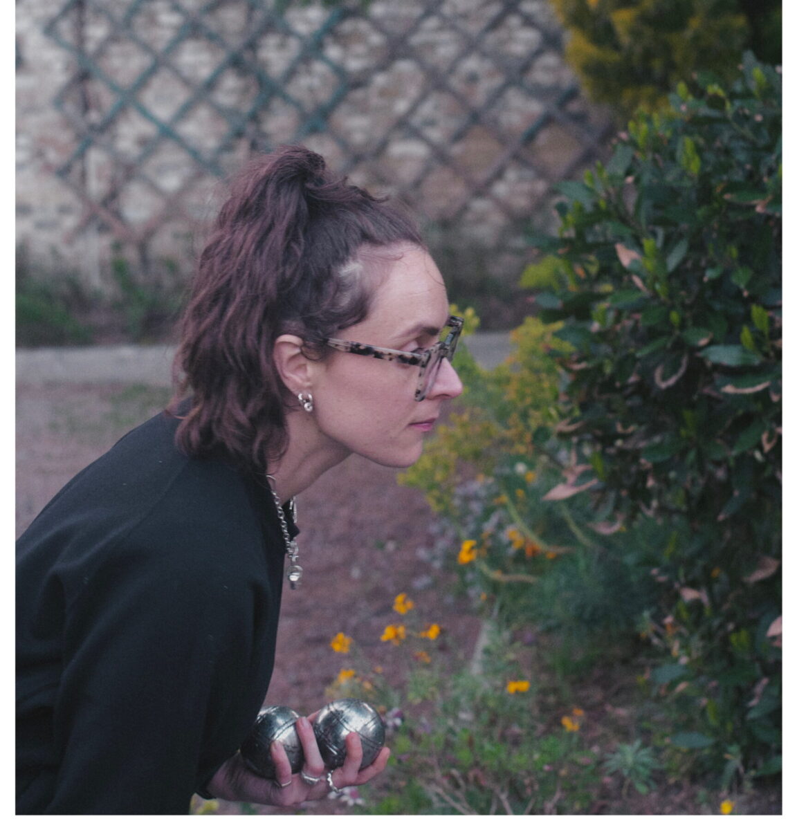 A woman leans over to smell the flowers groding in the hedgerow. There is a trellace fence behind her