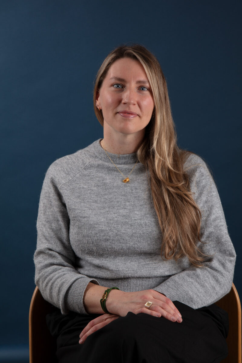 A jeweller sits on a chair looking at the camera with a blue background.