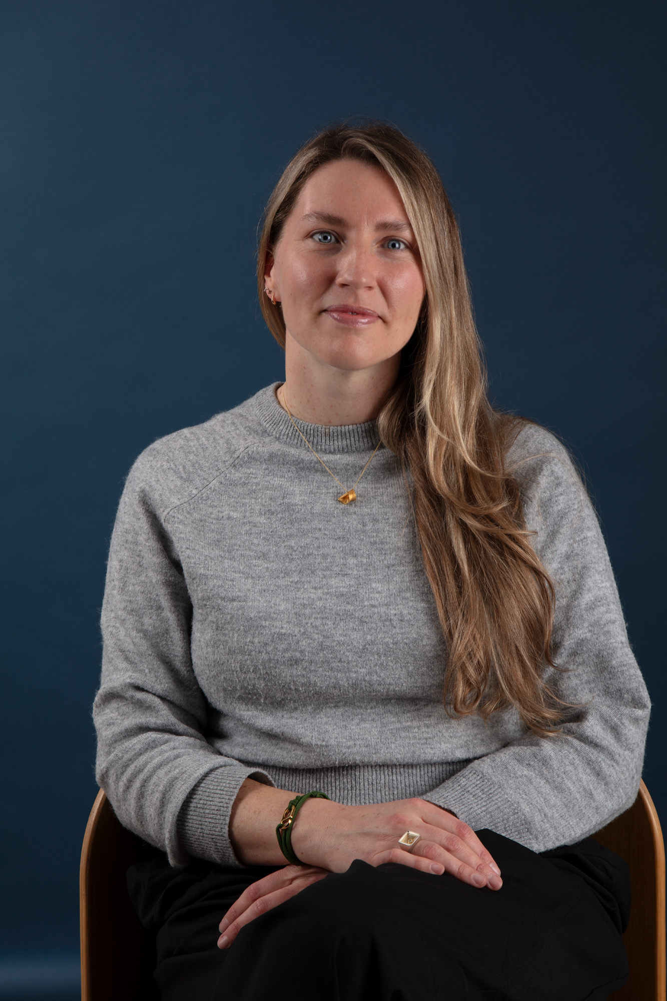 A jeweller sits on a chair looking at the camera with a blue background.