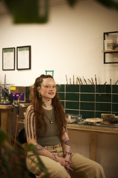 A tattooed woman sits on a chair surrounded by jewellery tools in a workshop