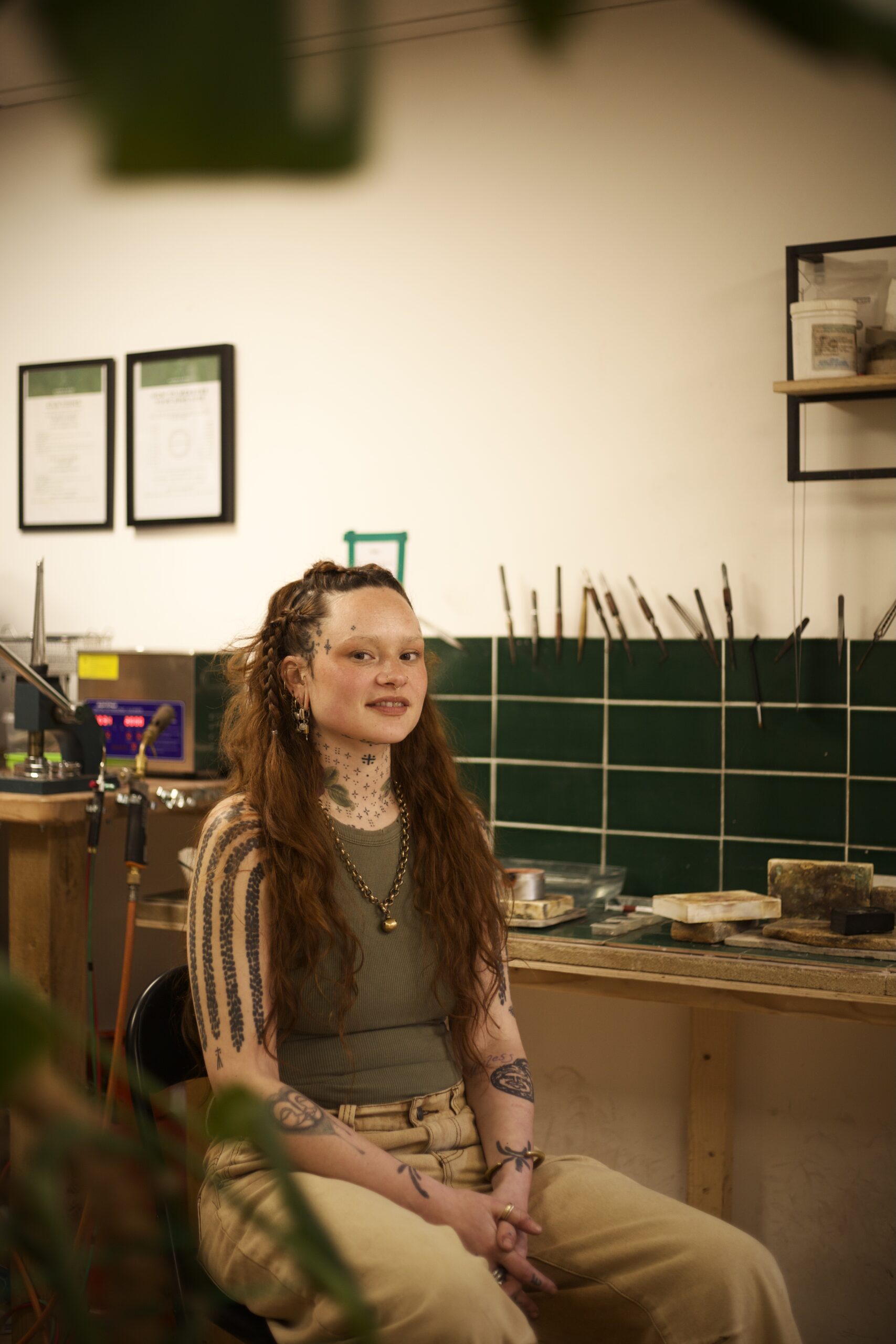 A tattooed woman sits on a chair surrounded by jewellery tools in a workshop