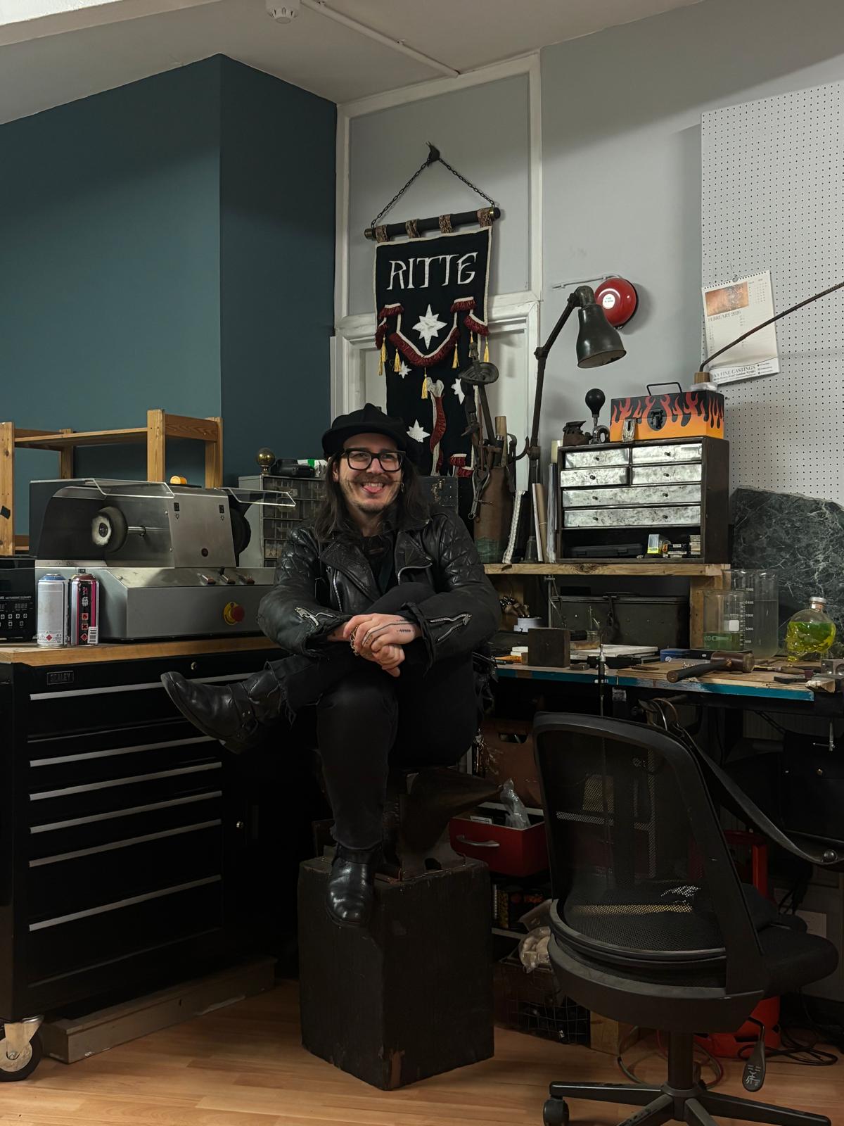 A man wearing a leather jacket and a flat cap perches on an anvil in a stocked tool workshop surrounded by tool cabinets and with a banner behind him.