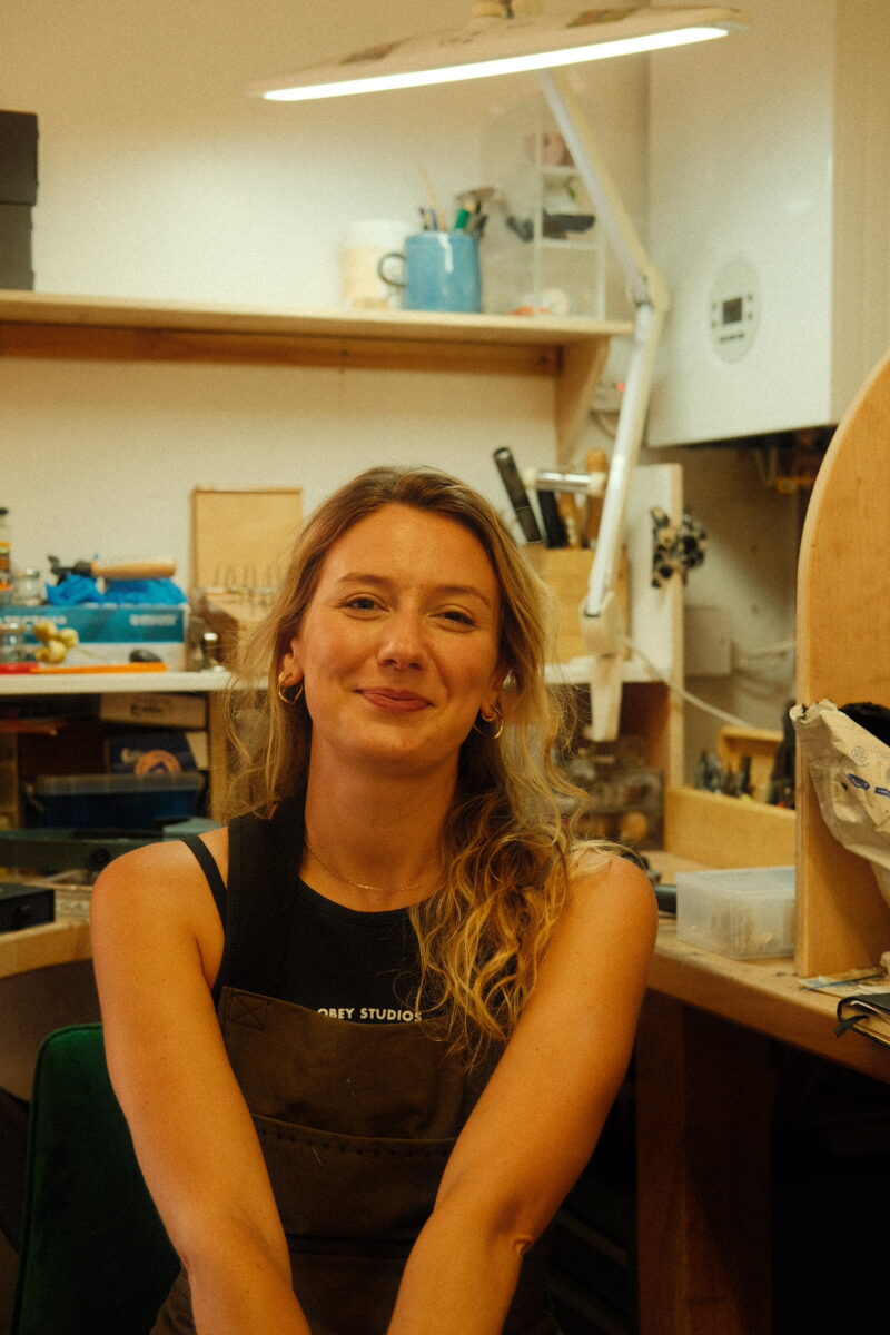 a smiling jeweller sits in front of a busy workbench